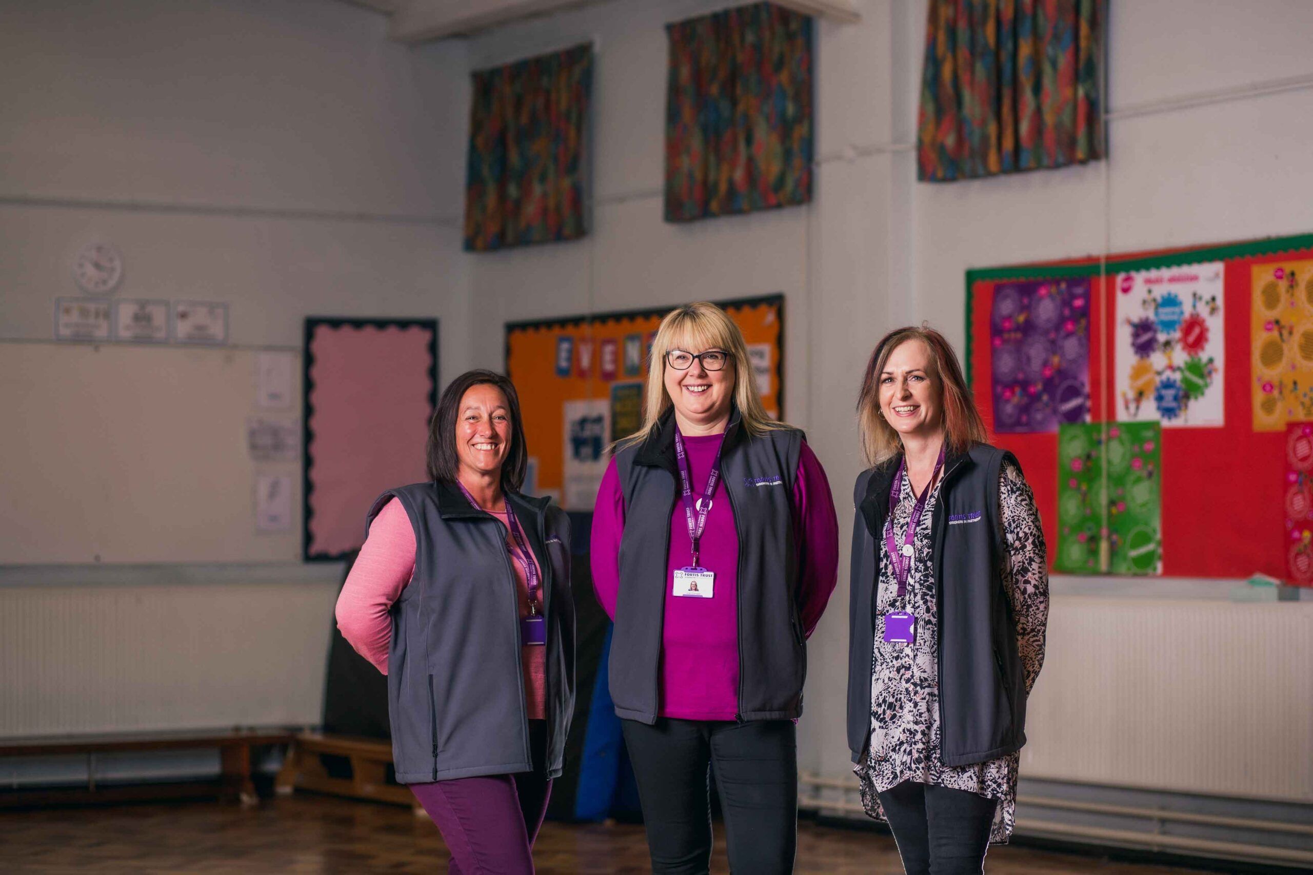Three members of the Fortis Trust Outreach Team. All three women are looking at the camera and are smiling. In the background is a school hall.