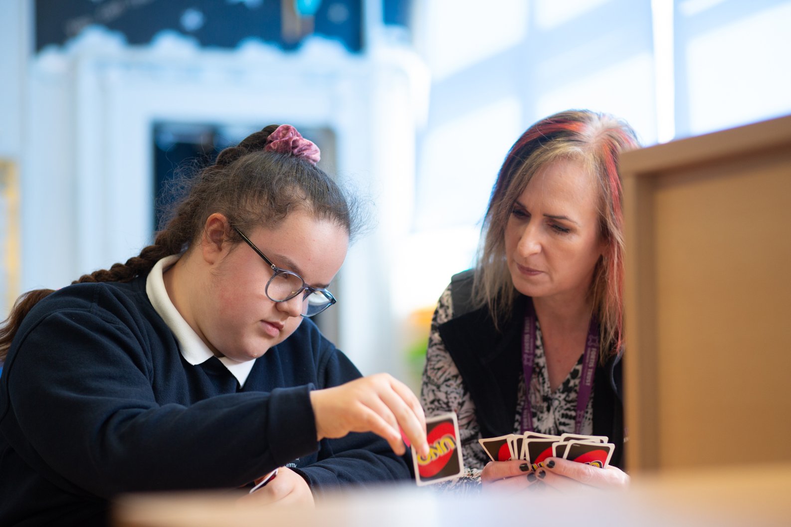 Student wearing glasses seated at a table, playing a card game with a member of Fortis Trust Outreach staff in a classroom or learning environment. The student places a card onto the table while the staff member holds several cards and observes. Shelving and bright windows are visible in the background.