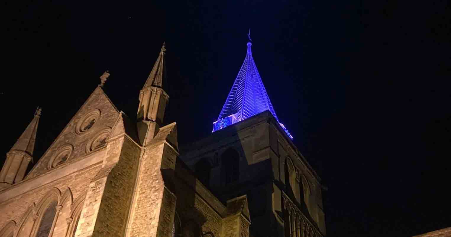 A nighttime view of Rochester Cathedral, illuminated by warm yellow lighting. Its tall central spire glows with bright purple, standing out against the dark sky. A crescent moon shines above the spire, and the building's detailed Gothic-style architecture is clearly visible.