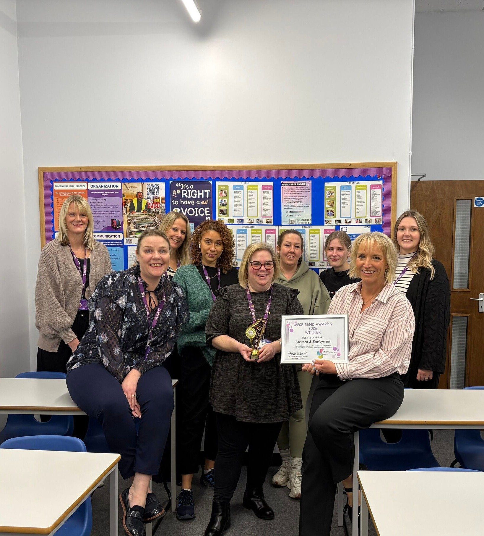 A group of staff standing and sitting in a classroom behind desks, with one person holding a certificate and another holding a trophy. A noticeboard with posters is visible behind them, and blue classroom chairs and tables are in the foreground.